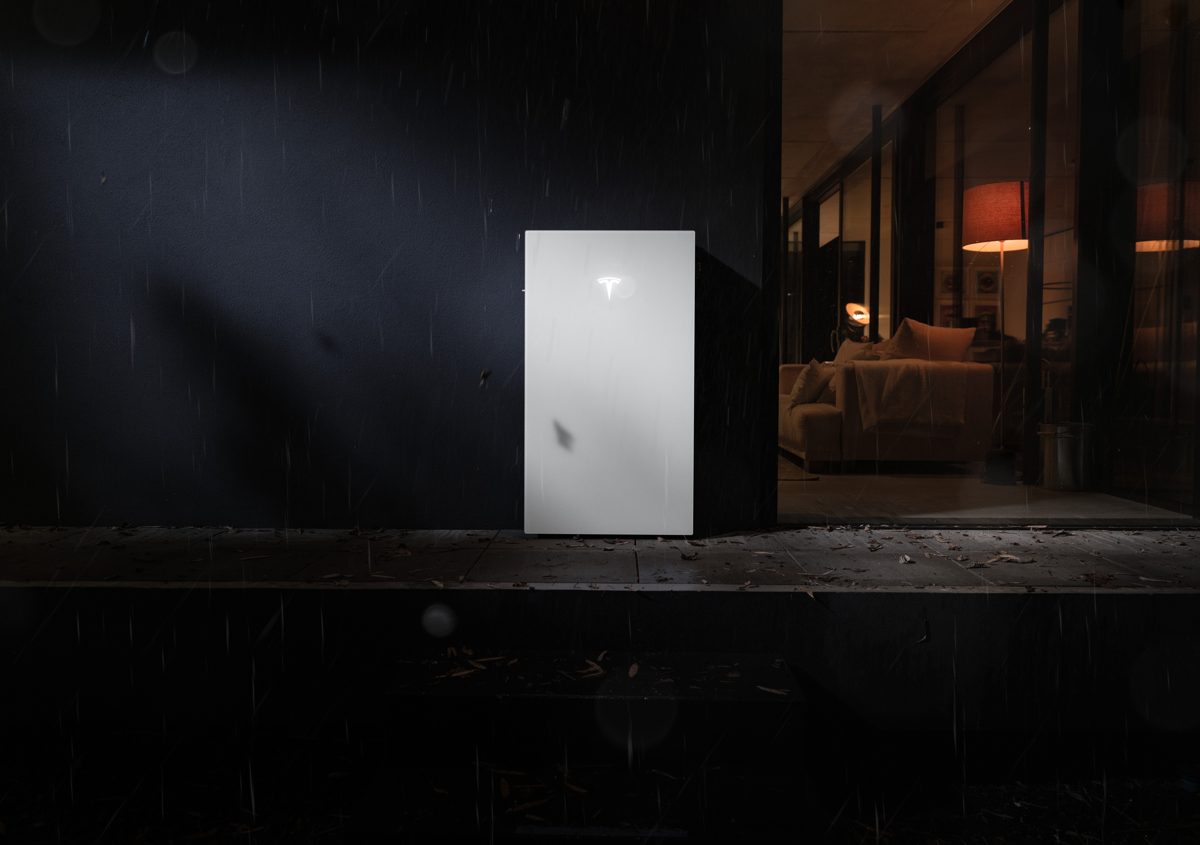 Tesla Powerwall 3P on an outdoor wall in the rain at dusk, with a warmly lit room behind